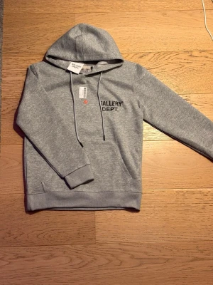 Grå hoodie från Gallery Dept. - Snygg grå hoodie från Gallery Dept. med svart tryck på bröstet och stor logga på ryggen. Klassisk känguruficka framtill, justerbar huva med snören och ribbade muddar. Materialet känns mjukt och bekvämt.                                        Skickas nästa dag📍.                    Kontakta för mer information🆔!