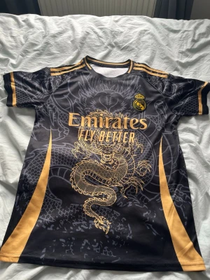 Real Madrid svart tränings t-shirt drake - Snygg svart Real Madrid tränings t-shirt från Adidas med guldiga detaljer och en stor drake på framsidan. Tröjan har klubbmärke, Emirates Fly Better-tryck och guldiga ränder på axlarna. Perfekt för fotboll eller gymmet. Priset kan sänkas men köpte den på stadium outlet för 800 kr och behöver snabba pengar så säljer den för 500 