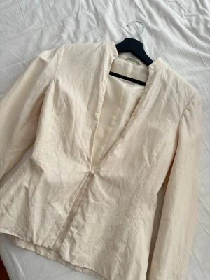 Krämvit tunn linne kavaj med broderi  - Så söt linne kavaj med  brodderi mönster på ärmarna. Kavajen har en enkel knäppning framtill. Säljer då den tyvärr inte kommer till användning. 45% linne 55% silk inner lager polyester. Stolek UK 10 men jag skulle säga att den passar strlk XXS-Xs