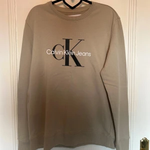 Beige Calvin Klein sweatshirt - Snygg beige sweatshirt från Calvin Klein Jeans med stor svart CK-logga och vit text på bröstet. Storlek L. 