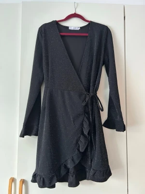 Svart omlottklänning med glitter - Svart omlottklänning från By Clara med långa ärmar och volangkant. Klänningen har ett subtilt glittrigt tyg och knyts i midjan för en snygg passform. Perfekt för dig som vill ha en chic look med lite extra shine.