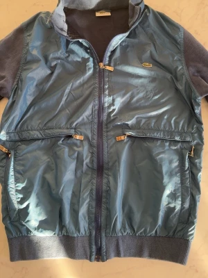 Blå vindjacka från Lacoste - Snygg blå vindjacka från Lacoste med glansig framsida och ribbade muddar. Jackan har två stora fickor med dragkedja och hel dragkedja framtill. Ärmarna är i mjukare tyg och jackan har en liten Lacoste-logga på bröstet.