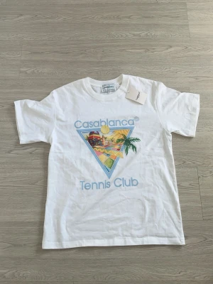Vit Casablanca Tennis Club t-shirt - Säljer en vit t-shirt från Casablanca med färgglatt tryck framtill som visar en solig tennis club-vibe, palm och landskap i triangelformad design. T-shirten har rund hals och korta ärmar. Materialet är mjuk bomull och trycket går i blått, gult, grönt och rött.