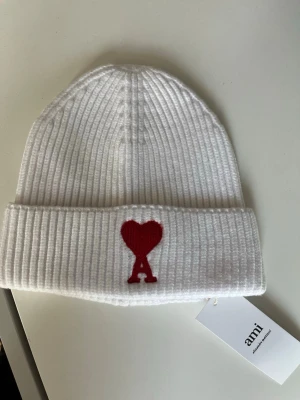 Vit ribbstickad mössa från Ami Paris  - Säljer en vit ribbstickad mössa från Ami Paris med uppvikt kant och röd broderad Ami de Coeur-logga framtill. Klassisk och stilren design som passar perfekt till kalla dagar. Mössan är i mjukt material och har en snygg passform.