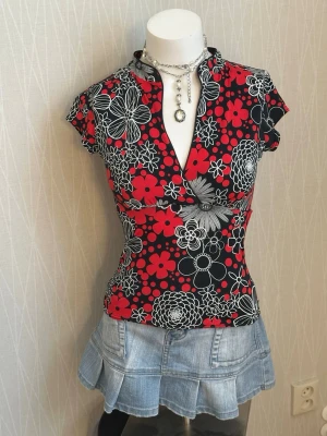 Blommig topp med v-ringning - Snygg topp med v-ringning och korta ärmar. Mönstret är blommigt i rött, svart och vitt, vilket ger en cool kontrast. Figurnära passform och stretchigt material som känns bekvämt. Perfekt för dig som vill sticka ut lite extra.
