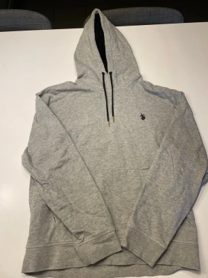 Grå hoodie från Polo Ralph Lauren - Snygg grå hoodie från Polo Ralph Lauren med svart broderad logga på bröstet. Klassisk känguruficka framtill, svarta dragsnören med gulddetaljer och mjukt material i bomullsblandning. Perfekt för en avslappnad och stilren look.