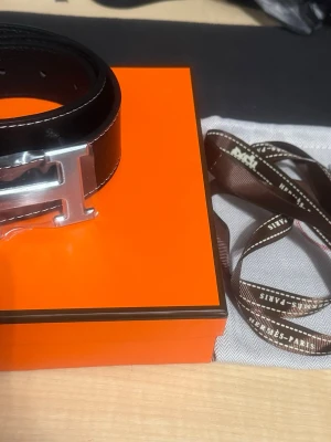 Svart skärp med silver H-spänne - Svart skärp i skinn med ett stort, blankt H-format spänne i silver. Skärpet har vita kontrastsömmar längs kanterna som ger en extra detalj. Perfekt för dig som gillar stilrena accessoarer med exklusiv känsla.