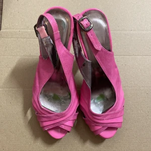 Rosa slingback pumps med öppen tå - Snygga rosa pumps med slingback-rem och öppen tå. Skorna har korslagda band framtill och justerbart spänne vid ankeln. Insidan är metallicfärgad och materialet ser ut att vara konstläder. Perfekta för dig som vill sticka ut med färg. Har slitage, lite repor och avskavt på insidan, se bilder.