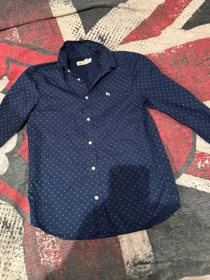 Mörkblå skjorta med småmönster - Mörkblå skjorta från Abercrombie & Fitch med små vitt mönster och broderad logga på bröstet. Skjortan har klassisk krage, knäppning framtill och långa ärmar. Perfekt för en stilren look.
