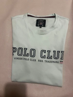 Vit t-shirt från Vinson Polo Club - Snygg vit t-shirt från Vinson Polo Club med tryckt text 'POLO CLUB' framtill. Klassisk rund halsringning och korta ärmar. Tillverkad i mjuk bomull som känns skön mot huden. Perfekt för en avslappnad och stilren look.