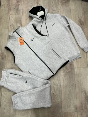 Grått Nike loungewear set - Mysigt loungewear set från Nike i ljusgrått med svarta detaljer. Setet består av en huvtröja med dragkedja, fickor och Nike-logga samt matchande byxor med resår i midjan och vid benslut. Perfekt för chill dagar hemma.