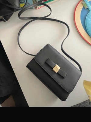 Svart axelväska med gulddetalj - Stilren svart axelväska med lång rem och dekorativ rosett på framsidan, prydd med en guldfärgad metallspänne. Väskan har en rektangulär form och är tillverkad i slätt syntetmaterial. Perfekt för dig som gillar minimalistisk design och snygga detaljer.