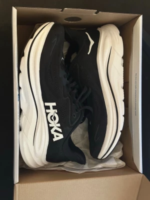Svarta Hoka sneakers med tjock sula - Snygga svarta sneakers från Hoka med vit tjock sula och tydlig logga på sidan. Skorna har meshmaterial för bra ventilation och klassisk snörning. Perfekta för dig som gillar sportig stil och komfort.