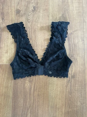 Svart spets-bh med breda axelband - Snygg svart bh i spets med breda axelband och dekorativt vågig kant. Bh:n har hakknäppning fram och är tillverkad i mjukt, stretchigt spetsmaterial. Perfekt för dig som gillar en feminin och bekväm stil.