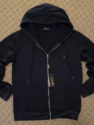 Marinblå hoodie från Polo Ralph Lauren - Snygg marinblå hoodie med dragkedja från Polo Ralph Lauren. Klassisk design med huva, dragsnören och kängurufickor. Liten röd logga broderad på bröstet. Tillverkad i mjukt bomullsmaterial för skön känsla.