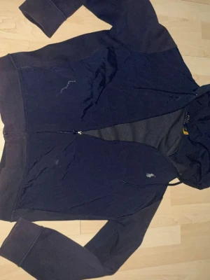 Mörkblå vindjacka från Polo Ralph Lauren - Snygg mörkblå vindjacka från Polo Ralph Lauren med dragkedja framtill och huva. Litet hål vid höger armbåge men inget som syns!