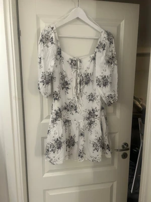 Vit blommig kortklänning med puffärm - Söt vit kortklänning med svart blommigt mönster och puffärmar. Klänningen har snörning framtill och volang nedtill, vilket ger en romantisk känsla. Perfekt för sommarens dagar och har en luftig passform. Materialet känns som bomull.