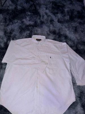 Vit randig skjorta från Ralph Lauren - Säljer en klassisk vit skjorta med tunna ljusgrå ränder från Ralph Lauren. Skjortan har lång ärm, knappar framtill och ikonisk broderad logga på bröstet. Perfekt för dig som gillar stilrena plagg med snygga detaljer.
