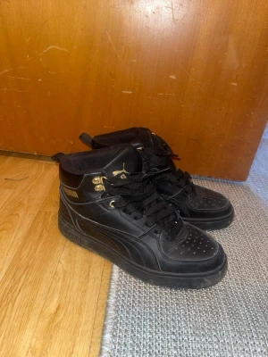 Svarta Puma high top sneakers - Snygga svarta high top sneakers från Puma med gulddetaljer och klassisk logga på sidan. Skorna har svart snörning, perforerad tå och är tillverkade i läderliknande material. Perfekta för dig som vill ha en stilren och sportig look.
