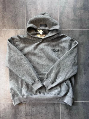 Grå hoodie Essentials Fear of God - Säljer en grå hoodie från Essentials Fear of God med svart logotyp på bröstet. Hoodien har en stor huva, ribbade muddar och en klassisk känguruficka. Materialet är mjukt och bekvämt, perfekt för en chill look. I storlek M fast oversized.