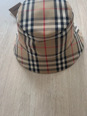 Burberry bucket hat med klassiskt rutmönster - Säljer en bucket hat från Burberry med ikoniskt beige, svart, vit och rött rutmönster. Hatten är tillverkad i slitstarkt tyg och har en rund form med bred brätte. Perfekt för att lyfta din streetstyle-look. OBS: Den här hatten är 100 % äkta och i nästintill nyskick. Etiketten är avtagen men finns självklart kvar och medföljer. Äktheten återspeglas tydligt i priset. Originalbox ingick tyvärr inte vid köpet, men digitalt kvitto finns som garanti.