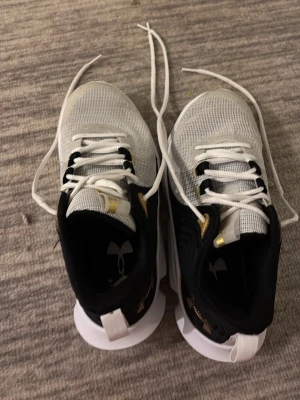 Under Armour vita och svarta sneakers - Snygga sneakers från Under Armour i vitt och svart med guldiga detaljer. Ovandelen är i mesh för bra andningsförmåga och sulan är vit med texten FLOW på sidan. Klassisk snörning och logga på hälen.