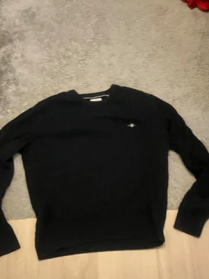Svart stickad tröja från GANT - Svart stickad tröja från GANT med klassiskt broderat logotyp på bröstet. Tröjan har rund halsringning och långa ärmar. Perfekt för dig som gillar stilrena och tidlösa plagg. Materialet är mjukt och bekvämt, med diskret kabelstickat mönster.