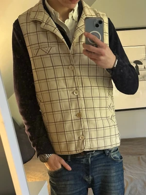Stenströms väst - Stilren beige väst från stenströms med brunt rutmönster och klassisk krage. Västen har knäppning framtill med fyra knappar och två fickor med dekorativa detaljer. Perfekt för att lyfta din outfit med en preppy touch. Storlek M köptes i butik för 2500 kr knappast använts 