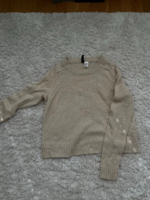 Beige stickad tröja från H&M - En enkel och stilren beige stickad tröja från H&M med rund halsringning och ribbade muddar vid ärmslut och nederkant. Perfekt för lager-på-lager och passar till det mesta i garderoben.