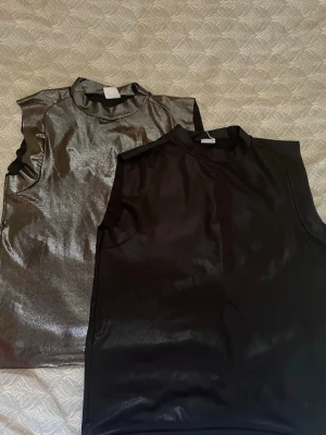 Glittriga metallic toppar Gina Tricot - Två glittriga metallic toppar från Gina Tricot, en i silver och en i svart. Båda är ärmlösa med hög hals och har en tight passform. Perfekta för att sticka ut och ge din outfit en extra edge.