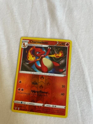 Charmeleon Pokémonkort - Charmeleon Pokémonkort med gul ram och färgstark illustration. Kortet har röda och orange detaljer, visar Charmeleon med eld i bakgrunden och har attacker som Slash och Raging Flames. Perfekt för samlare eller Pokémonfans.