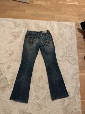 Lågmidjade bootcut jeans - Säljer ett par blå bootcut jeans från okänt märke med snygga slitningar och broderade detaljer på bakfickorna. 