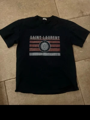 Svart Saint Laurent t-shirt med tryck - Cool svart t-shirt från Saint Laurent med ett stort tryck framtill i rött och vitt. Trycket har texten 'Saint Laurent' och ett emblem med SL i mitten. Klassisk passform och rund hals, perfekt till jeans eller shorts.