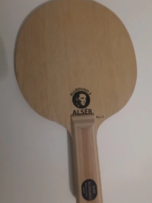 Bordtennisracket AL SÉR Allround - Säljer ett klassiskt bordtennisracket från ALSÉR, modell Allround 5. Racketen är i trä med en ljusbrun färg och har en rektangulär handtag. Perfekt för dig som gillar pingis och vill ha ett stabilt racket att spela med. Första riktiga stomme