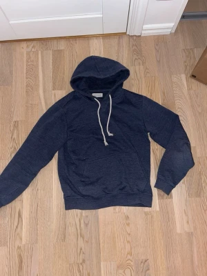 Mörkblå hoodie från Pier One - Mörkblå hoodie från Pier One med klassisk huva och vita snören. Hoodien har en stor magficka och ribbade muddar vid ärmslut och nederkant. Perfekt för en chill och avslappnad stil.