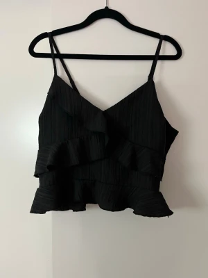 Svart volang topp med smala band - Snygg svart topp med smala axelband och volangdetaljer framtill och nedtill. Toppen har en omlott-look vid bysten och är croppad i modellen. Perfekt för dig som gillar trendiga och stilrena plagg. Toppen har en dragkedja på sidan