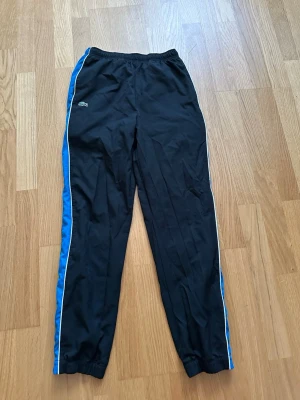 Svarta mjukisbyxor från Lacoste - Svarta mjukisbyxor från Lacoste med blåa sidor och vit piping längs benen. Byxorna har resår i midjan och vid benslut, samt Lacoste-logga och broderad krokodil på ena benet. Perfekta för en sportig och avslappnad stil.