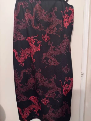 Svart klänning med röda drakar - Säljer en svart klänning med tunna axelband och ett coolt rött drakmönster över hela plagget. Klänningen har en rak passform och slutar ungefär vid knäna. Perfekt för dig som vill sticka ut med en unik och edgy stil.