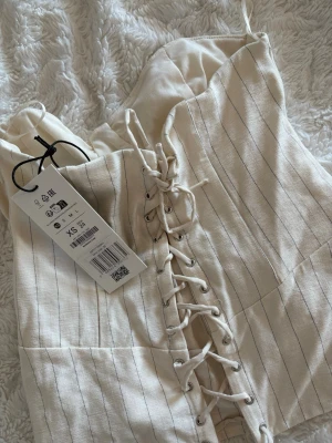 Randig korsettopp - Superfin beige korsettopp från Bershka med tunna svarta ränder och snygg snörning framtill. Toppen är axelbandslös och har en figurnära passform med markerade kupor. Perfekt att styla med jeans eller kjol för en trendig look.