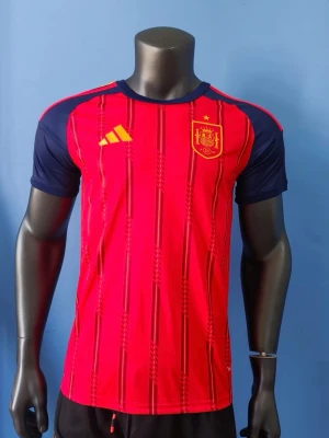 Spaniens matchtröja Adidas röd/blå - Säljer en officiell matchtröja från Spaniens landslag, designad av Adidas. Tröjan är röd med vertikala mönster, har mörkblå ärmar och gula detaljer. Landslagets emblem och Adidas-logga på bröstet. Tillverkad i lätt och ventilerande material, perfekt för fotboll.