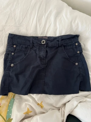 Mörkblå kortkjol med fickor - Snygg mörkblå kortkjol med flera fickor och dekorativa knappar framtill och baktill. Kjolen har bälteshällor och coola spännen bak. Perfekt för dig som gillar en casual och trendig stil.