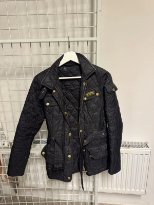 Svart quiltad jacka från Barbour - Snygg svart quiltad jacka från Barbour med guldiga knappar och dragkedja. Jackan har flera fickor framtill, klassisk krage och en figurnära passform. Perfekt för höst och vår när du vill ha en stilren look.