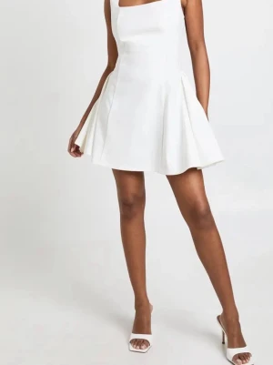 Vit kort klänning med breda axelband - Stilren vit kort klänning med breda axelband och fyrkantig urringning. Klänningen har en utsvängd kjol och är ärmlös, vilket ger en fräsch och modern look. Perfekt för sommarens alla tillfällen.