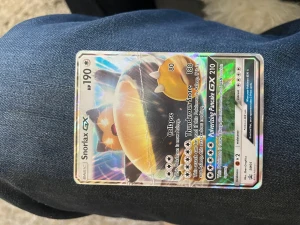 Snorlax GX Pokémonkort holografiskt - Snorlax GX Pokémonkort med holografisk yta. Kortet har färger i blått, gult och vitt med Snorlax-motiv och flera attacker tryckta på framsidan. Baksidan har klassisk Pokéball-design. Perfekt för samlare eller Pokémonfans.