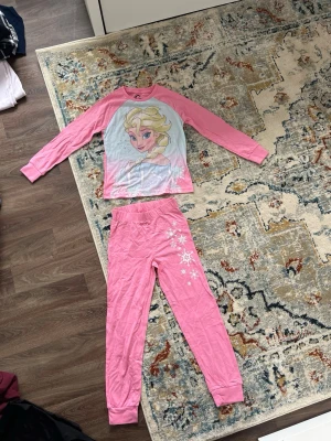 Rosa Frost pyjamas med Elsa-motiv - Superfin rosa pyjamas med långärmad tröja och byxor, prydd med stort Elsa-tryck från Frost på tröjan och snöflingor på byxbenet. Perfekt för alla som älskar Disney och vill mysa i något bekvämt. Mjukt material och stretchig midja.