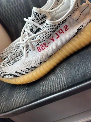 Adidas Yeezy Boost 350 Zebra - Säljer ett par Adidas Yeezy Boost 350 Zebra med stickad ovandel i vitt och svart mönster, röda 'SPLY-350' detaljer på sidan och en gulaktig Boost-sula. Skorna har klassisk låg profil och är tillverkade i mesh och syntetmaterial. Perfekt för dig som gillar streetwear och sneakers med unik design.