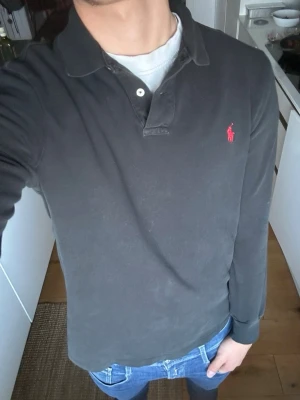 Svart långärmad pikétröja Ralph Lauren - Svart långärmad pikétröja från Ralph Lauren med klassisk krage och knäppning vid halsen. Tröjan har det ikoniska röda broderade logotypen på bröstet och ribbade muddar vid ärmsluten. Perfekt för en stilren och avslappnad look.