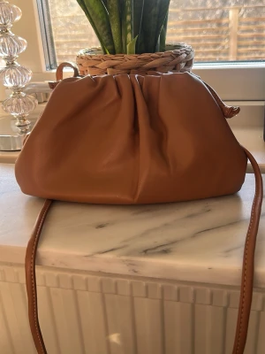 Brun axelväska från raffinato  - Snygg brun axelväska i mjukt fejkläder med rynkad design och lång axelrem. Väskan har en modern, puffig form och stängs upptill. Perfekt för dig som vill ha en trendig och stilren accessoar till din outfit. Originalpris- 500