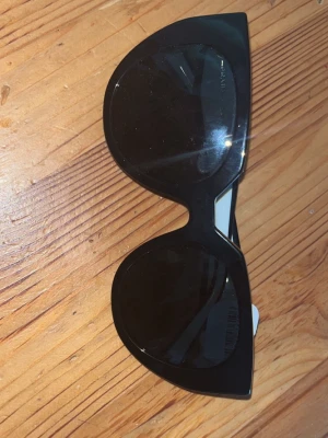 Svarta cat eye solglasögon från Prada - Snygga svarta solglasögon från Prada med cat eye-formade bågar och breda vita skalmar där Prada-loggan syns tydligt. Glasen är mörka och bågarna är tillverkade i plast. Perfekt accessoar för en trendig look.