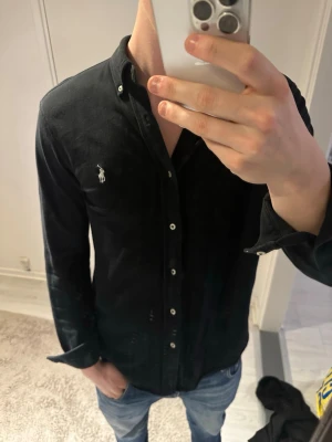 Svart skjorta från Ralph Lauren - Klassisk svart skjorta från Ralph Lauren med vit broderad logga på bröstet. Skjortan har lång ärm, knappar framtill och krage med knappar. Tillverkad i mjuk bomull och har en stilren, tidlös design.